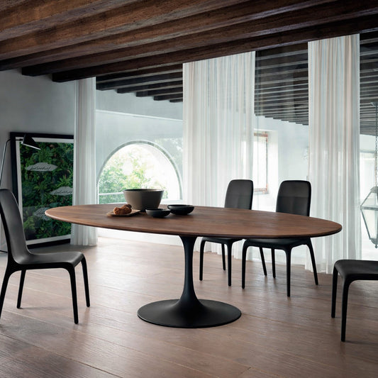Oval wooden dining table with black pedestal base in a modern architectural interior with exposed wooden beams, sheer curtains and arched window, styled with black chairs