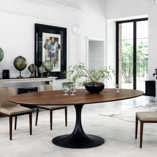 Oval wooden dining table with black pedestal base in a modern classic dining room, styled with upholstered chairs, fireplace, decorative objects and large windows