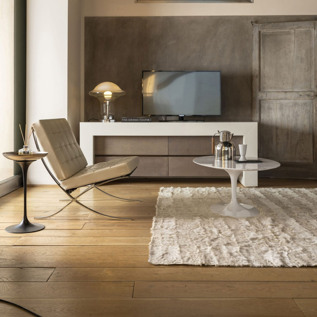 Barcelona Chair upholstered in beige tufted leather with polished chrome frame beside a white Tulip side table with a grey marble top, styled in a contemporary minimalist living room with warm wood flooring and neutral décor