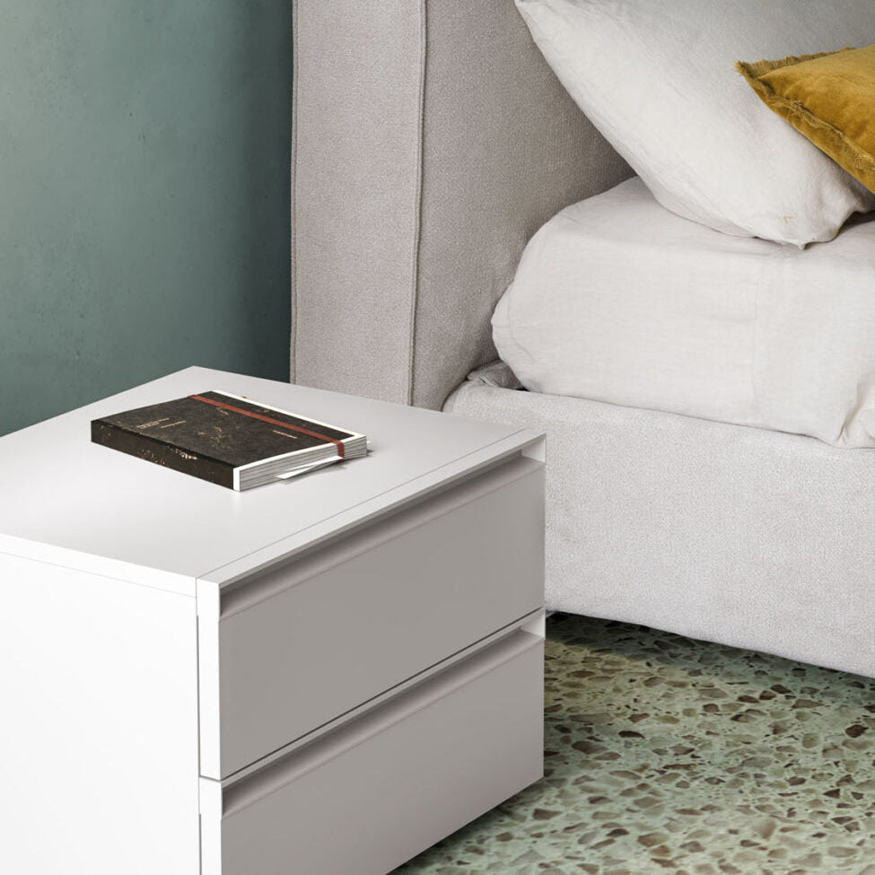 Gray upholstered bed with white bedding and a nightstand with a book against a gray wall.