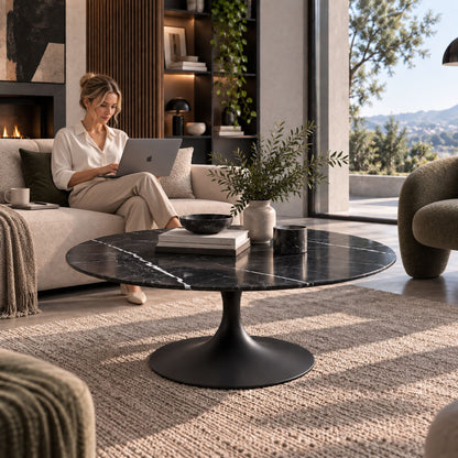 Woman working on a laptop beside a black marble-effect oval Tulip coffee table with a black pedestal base in a contemporary luxury living room.