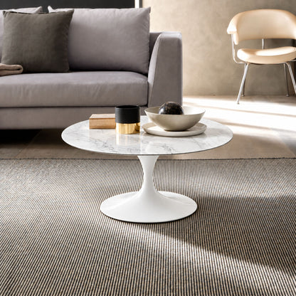 Carrara Elegance Tulip coffee table featuring a polished Carrara marble tabletop with subtle grey veining and a glossy white tulip pedestal base. Styled in a contemporary living room with a grey sofa, modern accent chair, textured rug, and decorative accessories, showcasing timeless Italian design for modern, Scandinavian, and mid-century interiors