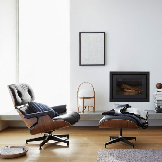 Eames-style lounge chair and matching ottoman with walnut veneer shell, black leather upholstery and black swivel base in a modern living room beside a contemporary fireplace.