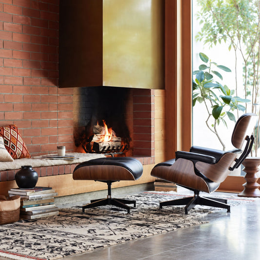 Eames-style lounge chair and matching ottoman featuring walnut veneer shell, black leather upholstery and a black swivel base, displayed in a contemporary living room beside a fireplace.
