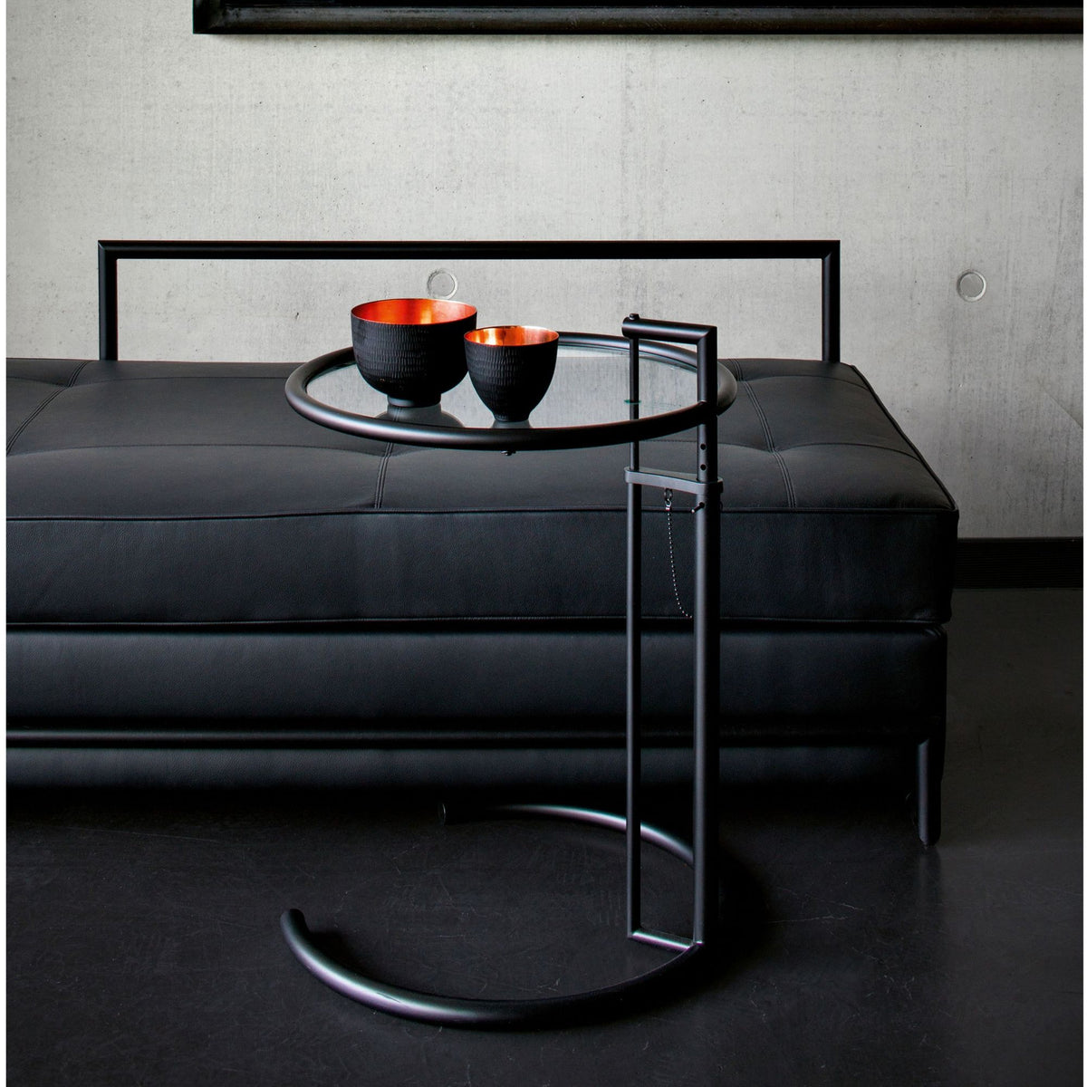 Eileen Gray E1027 black adjustable side table with a clear glass top and matte black tubular steel frame, styled beside a contemporary black leather sofa with decorative bowls, showcasing an iconic Bauhaus designer accent table for modern luxury living rooms.