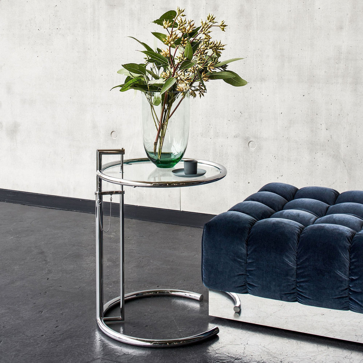 Eileen Gray E1027 chrome adjustable side table with a clear glass top and polished tubular steel frame, styled beside a contemporary blue upholstered bench with decorative vase, combining iconic Bauhaus design with modern luxury interiors
