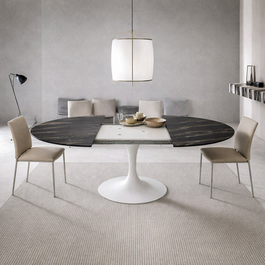 Extendable round tulip dining table with black ceramic top and white pedestal base, shown open with extension leaves, paired with beige upholstered chairs in a modern minimalist dining room.