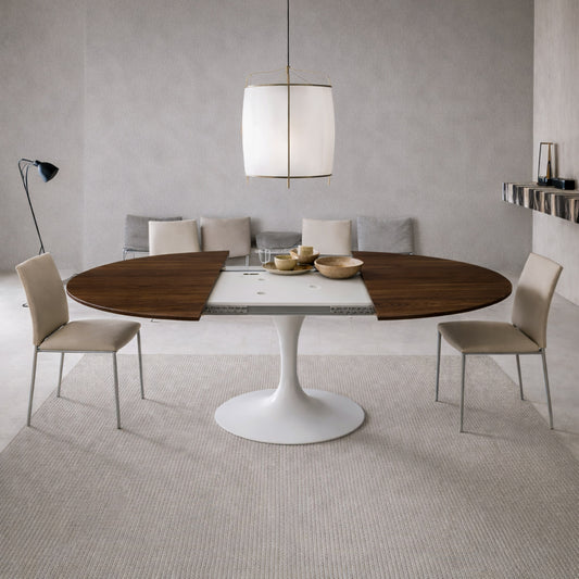 Extendable round tulip dining table with walnut wood top and white pedestal base, shown open with extension leaves in a modern minimalist dining room with beige upholstered chairs and neutral tones.