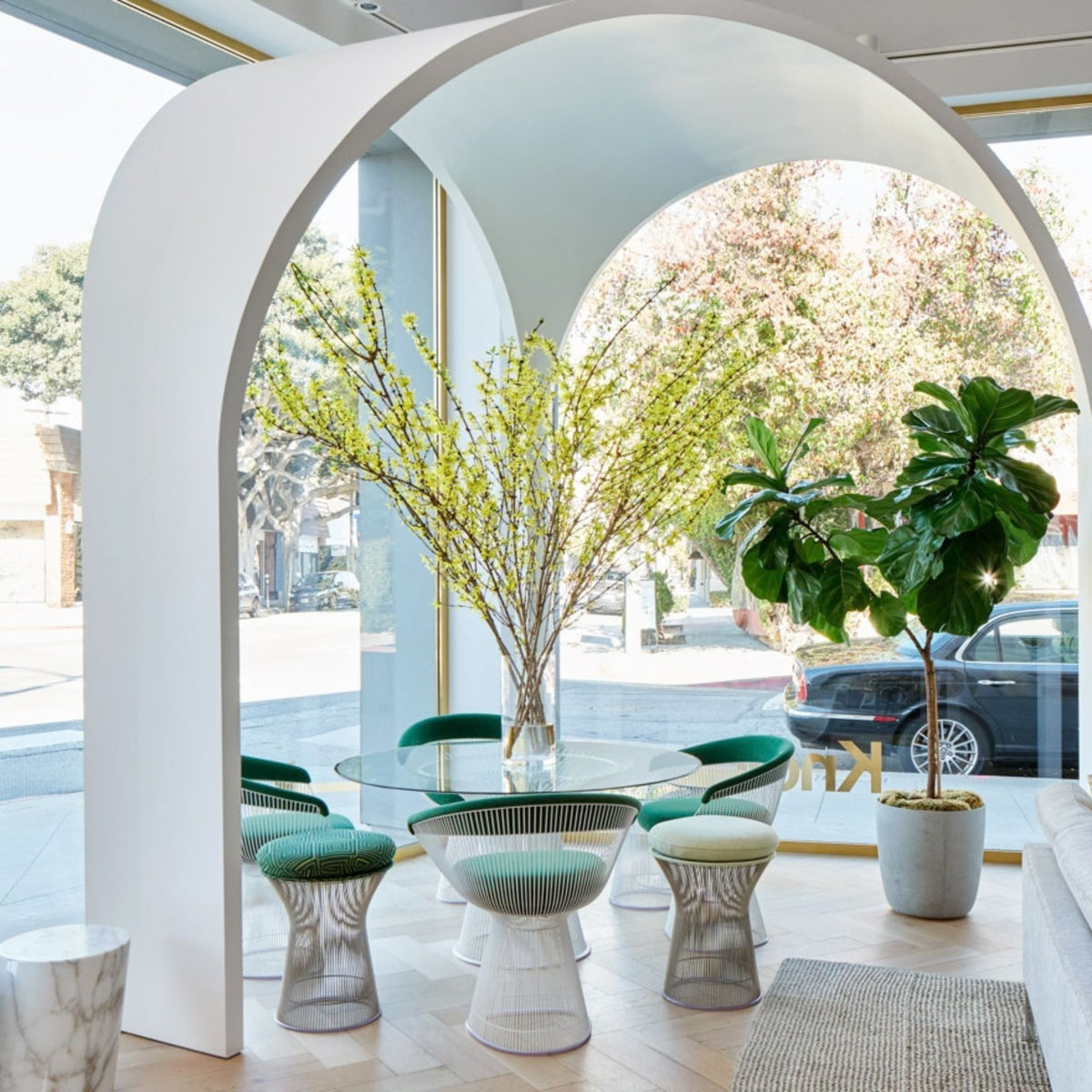 Modern dining space featuring a round glass dining table surrounded by green upholstered Platner wire dining chairs and matching Platner stools with polished steel rod bases beneath a sculptural white arch.