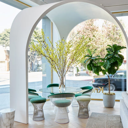 Modern dining space featuring a round glass dining table surrounded by green upholstered Platner wire dining chairs and matching Platner stools with polished steel rod bases beneath a sculptural white arch.
