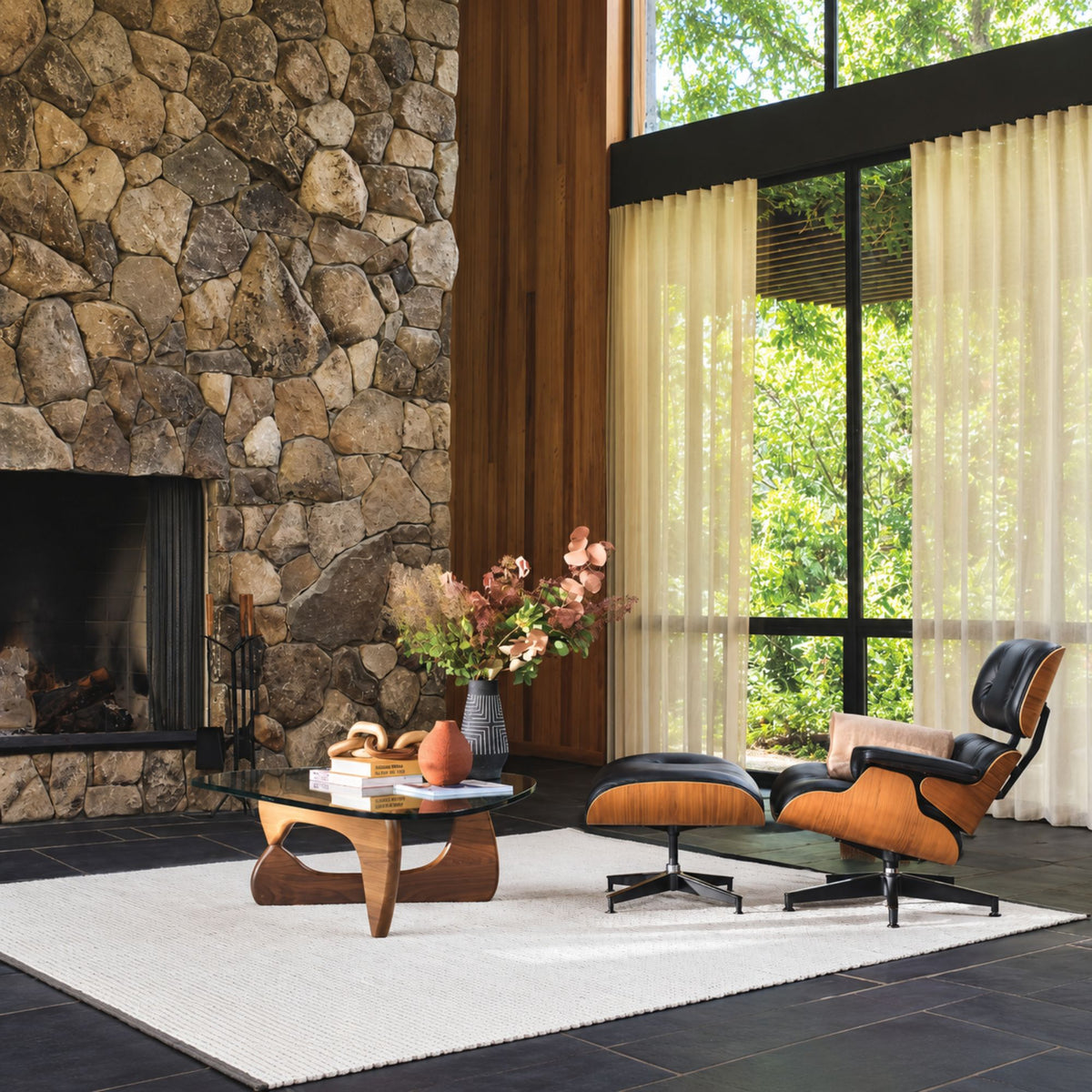 Isamu Noguchi glass coffee table with a sculptural walnut wood base, styled with a black and walnut Eames Lounge Chair and Ottoman in a luxury living room featuring a natural stone fireplace, showcasing iconic mid-century modern designer furniture.