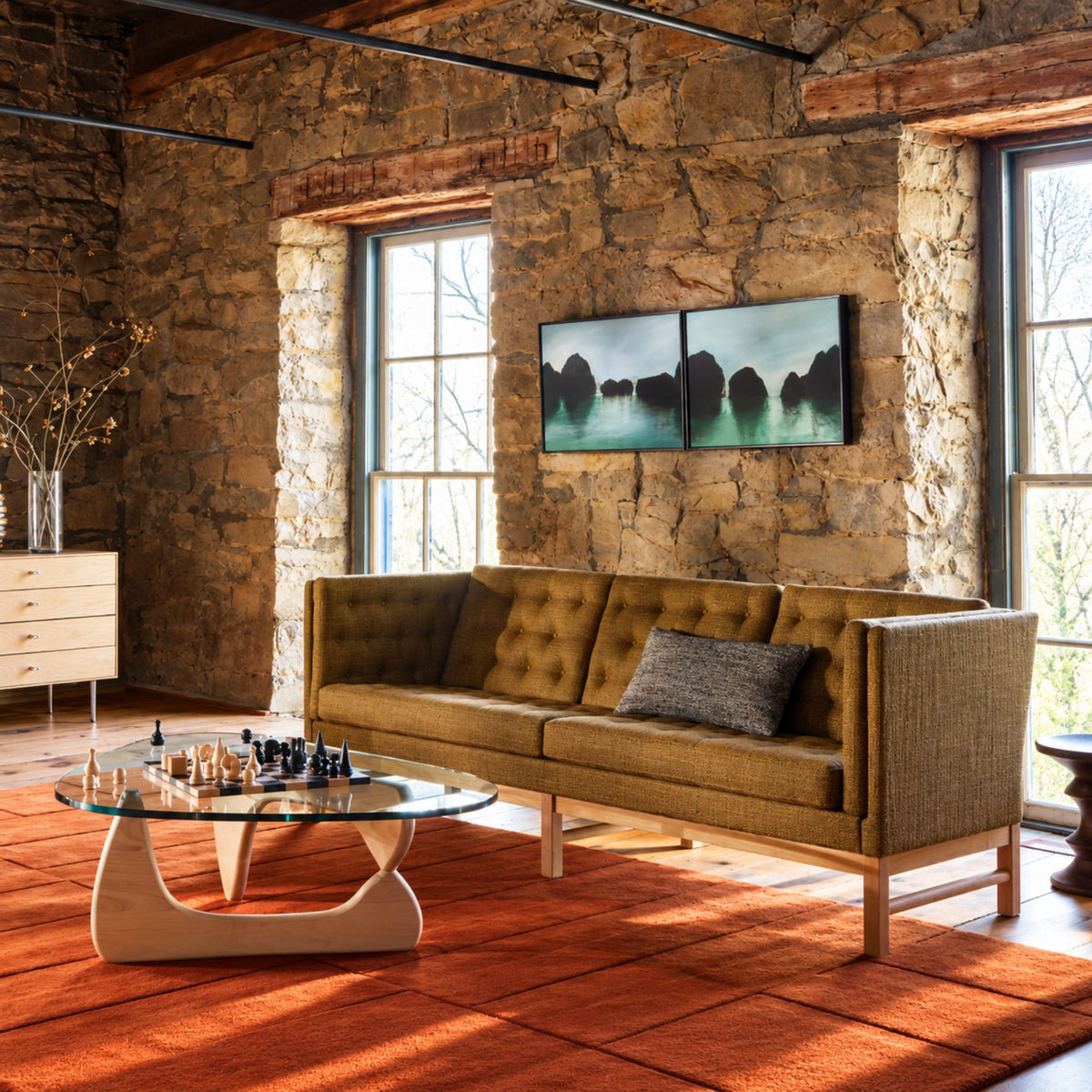 Isamu Noguchi glass coffee table with a sculptural solid wood base and organically shaped glass top, styled in a rustic modern living room with a tufted sofa, chess set, and warm natural lighting, showcasing an iconic mid-century modern designer coffee table