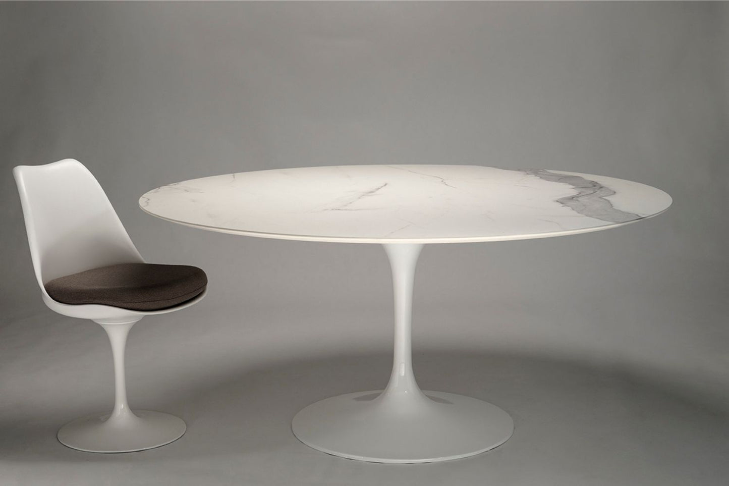 Oval ceramic tulip dining table with white marble-effect top and white pedestal base, paired with a single matching chair on a neutral gray background