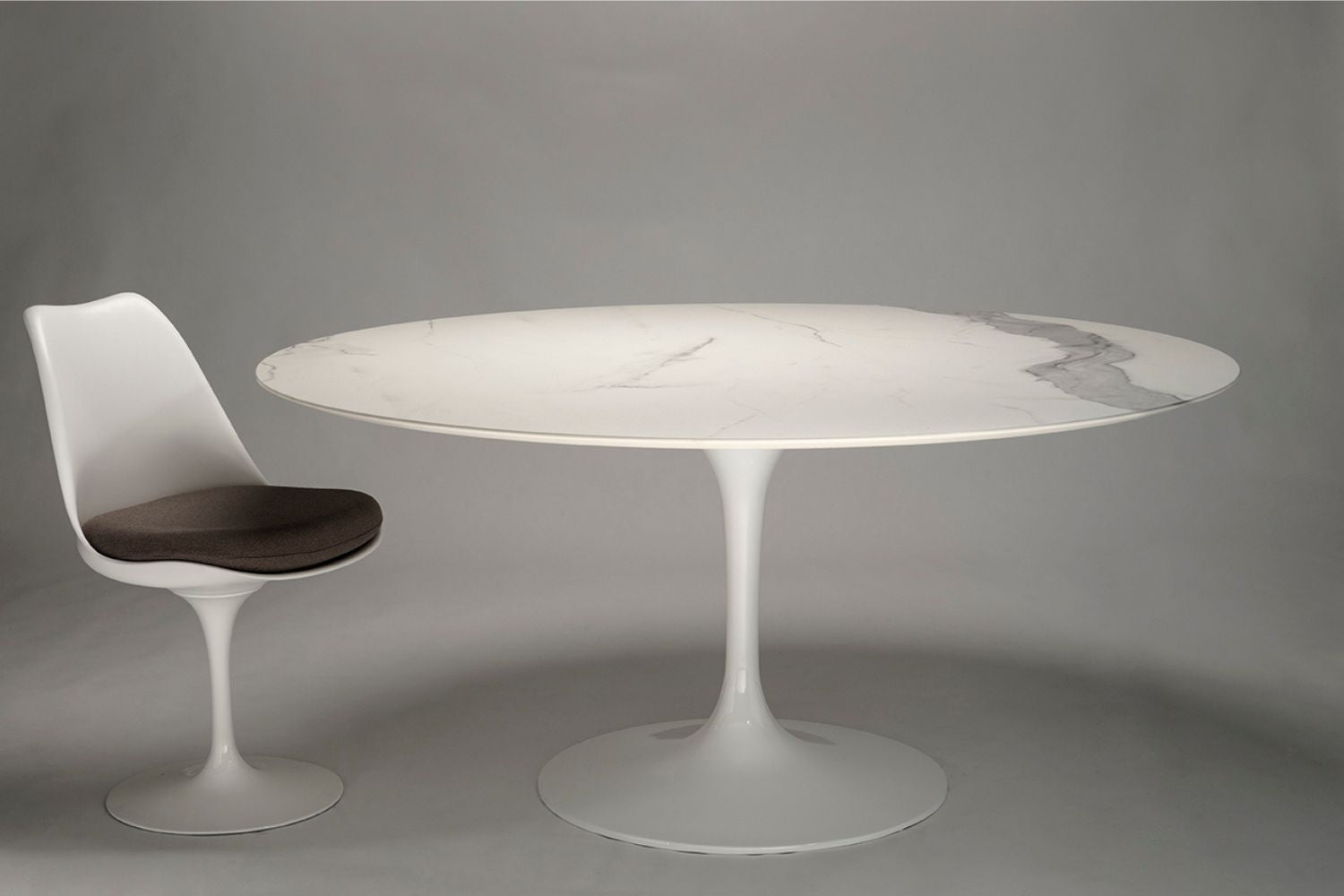 Oval ceramic tulip dining table with white marble-effect top and white pedestal base, paired with a single matching chair on a neutral gray background