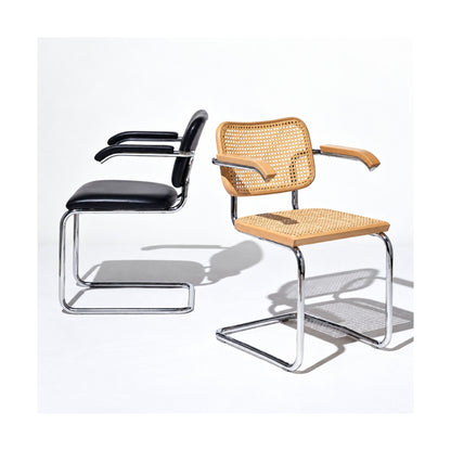 Marcel Breuer Cesca armchair collection featuring a natural cane armchair with wooden armrests beside a black upholstered leather cantilever armchair, both with polished chrome tubular steel frames on a white background.