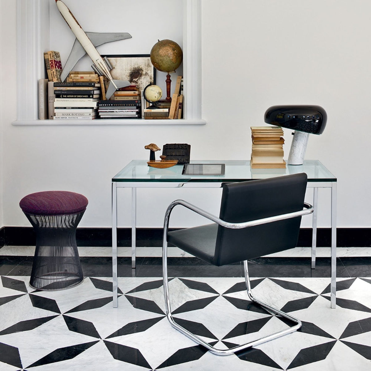 Modern home office featuring a purple upholstered Platner wire stool, a black leather cantilever desk chair with a chrome frame, and a glass-top desk in a bright contemporary workspace