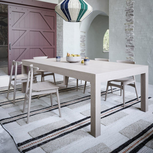 Modern light oak rectangular dining table with thick square legs, styled with upholstered dining chairs in a bright contemporary dining room featuring a statement pendant light and large windows