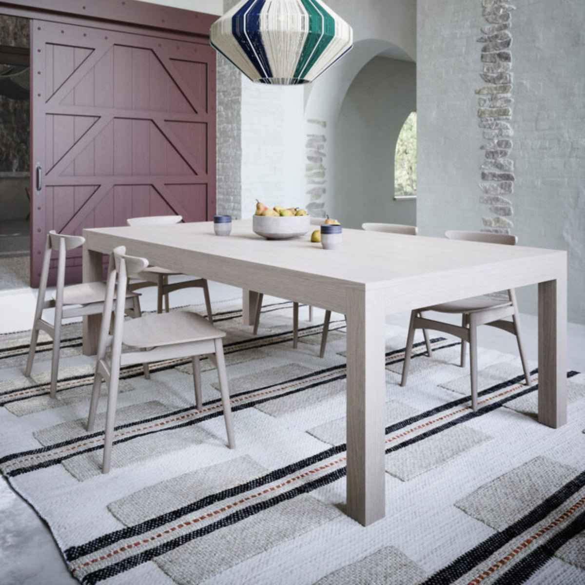 Modern light oak rectangular dining table with thick square legs, styled with upholstered dining chairs in a bright contemporary dining room featuring a statement pendant light and large windows