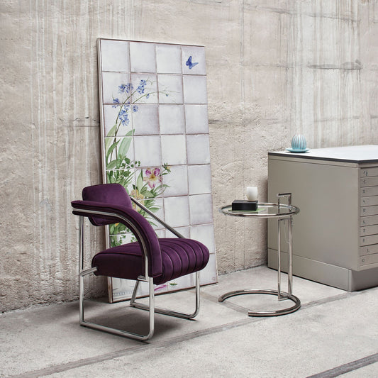 Modern dining armchair upholstered in luxurious purple velvet with a ribbed padded seat, curved upholstered armrest and polished chrome tubular sled base, styled in a contemporary interior alongside a chrome side table, combining elegant Italian-inspired design with everyday comfort.