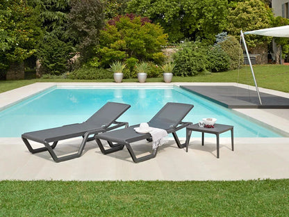 Two modern outdoor sun loungers with a matching side table beside a swimming pool in a landscaped garden.