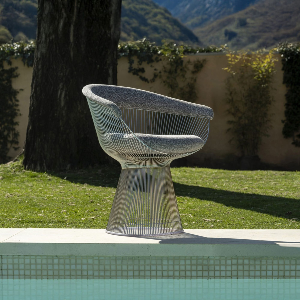 Outdoor wire-frame lounge chair with a sculptural stainless steel rod base and grey upholstered seat cushion, inspired by Warren Platner's iconic mid-century modern design, positioned beside a swimming pool.