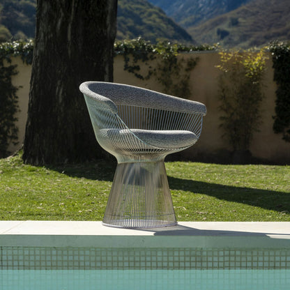 Outdoor wire-frame lounge chair with a sculptural stainless steel rod base and grey upholstered seat cushion, inspired by Warren Platner's iconic mid-century modern design, positioned beside a swimming pool.