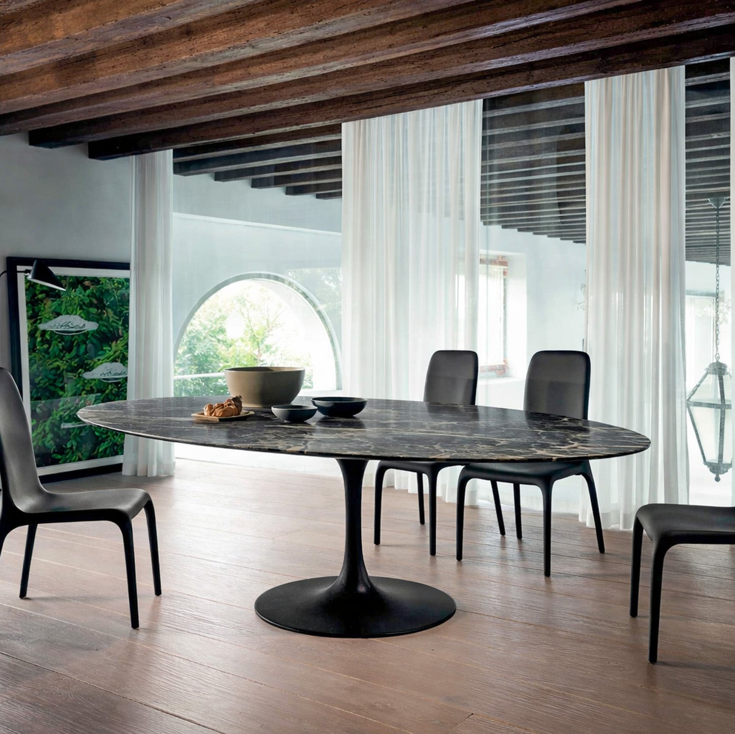 Oval dark marble dining table Saarinen style with black pedestal base in a modern architectural interior with exposed wooden beams and arched window, styled with black chairs