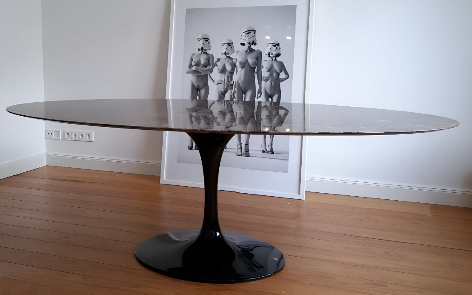 Oval tulip dining table with black marble-effect top and black pedestal base in a minimal interior setting with wooden flooring and framed artwork