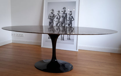Oval tulip dining table with black marble-effect top and black pedestal base in a minimal interior setting with wooden flooring and framed artwork