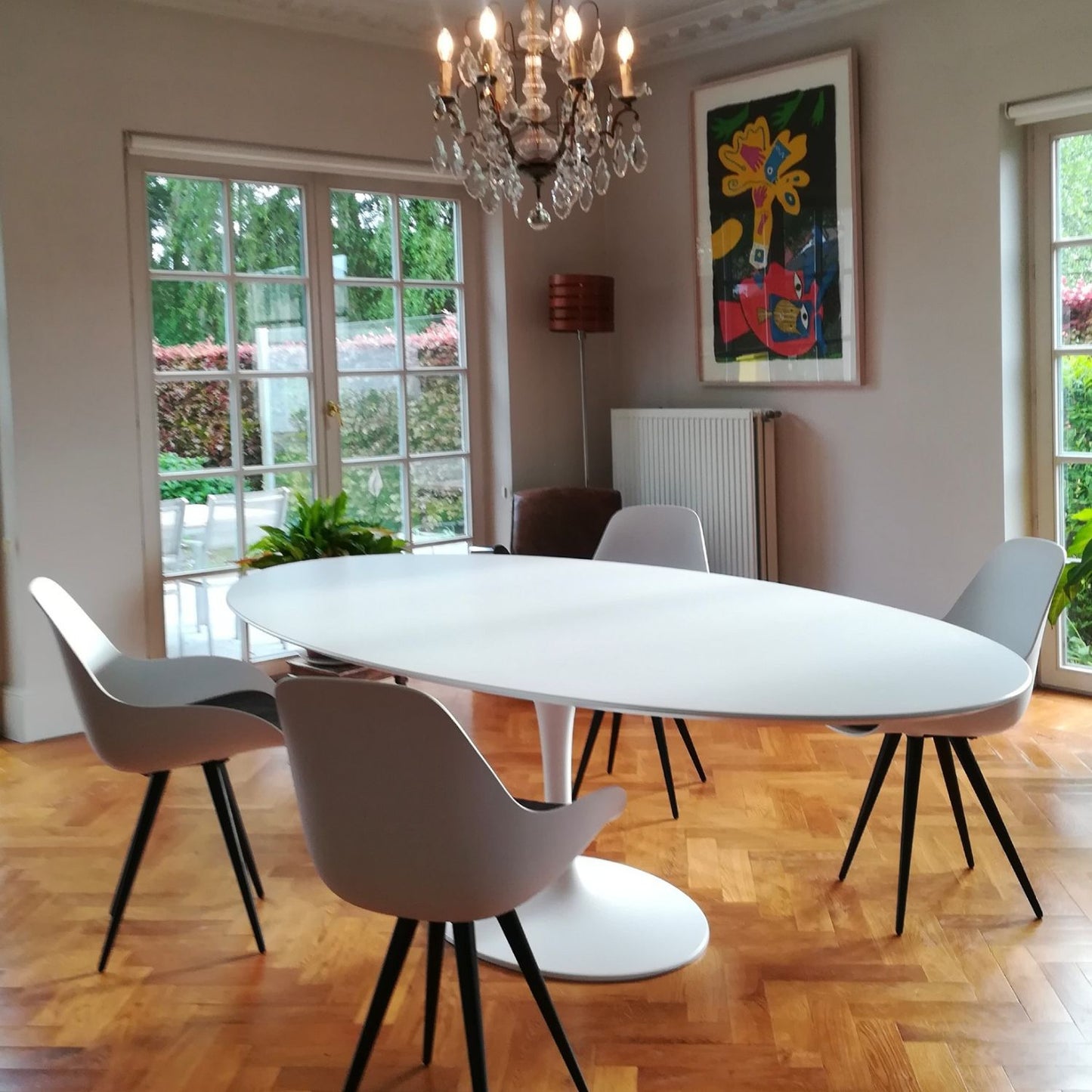 Oval tulip dining table in Saarinen style with white laminate top and white pedestal base, styled in a modern dining room with upholstered chairs, chandelier and wall art