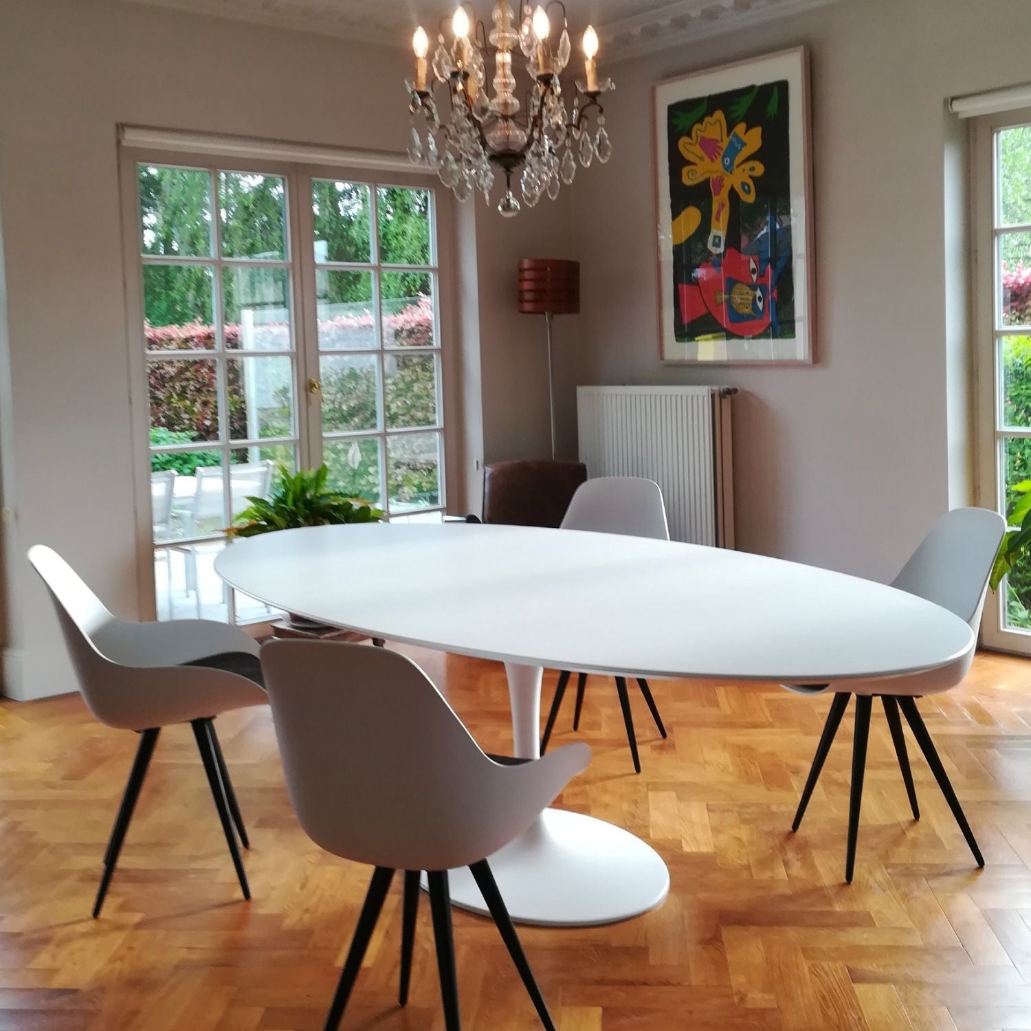 Oval tulip dining table in Saarinen style with white laminate top and white pedestal base, styled in a modern dining room with upholstered chairs, chandelier and wall art
