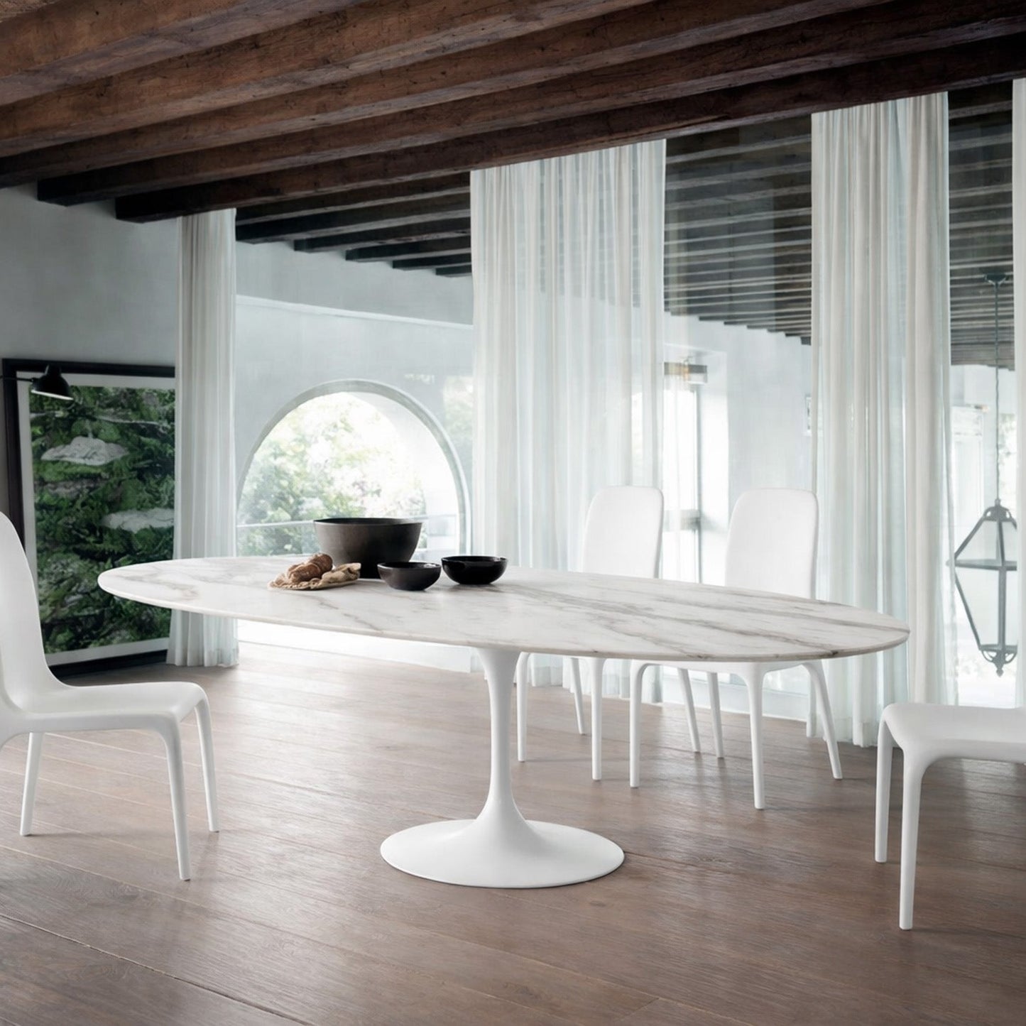 Oval white marble dining table Saarinen style with white pedestal base in a modern architectural dining room with exposed wooden beams, arched window and sheer curtains, styled with white chairs
