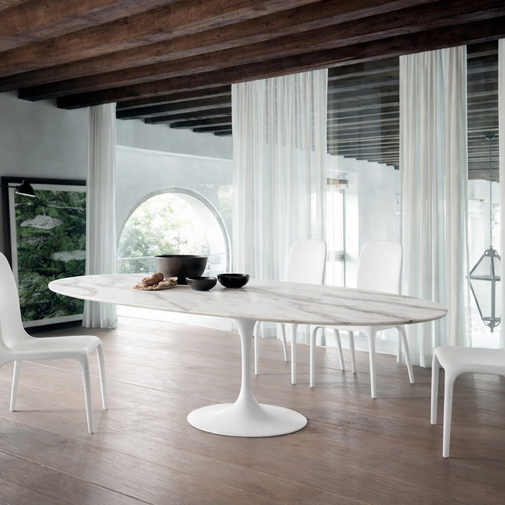 Oval white marble dining table Saarinen style with white pedestal base in a modern architectural dining room with exposed wooden beams, arched window and sheer curtains, styled with white chairs