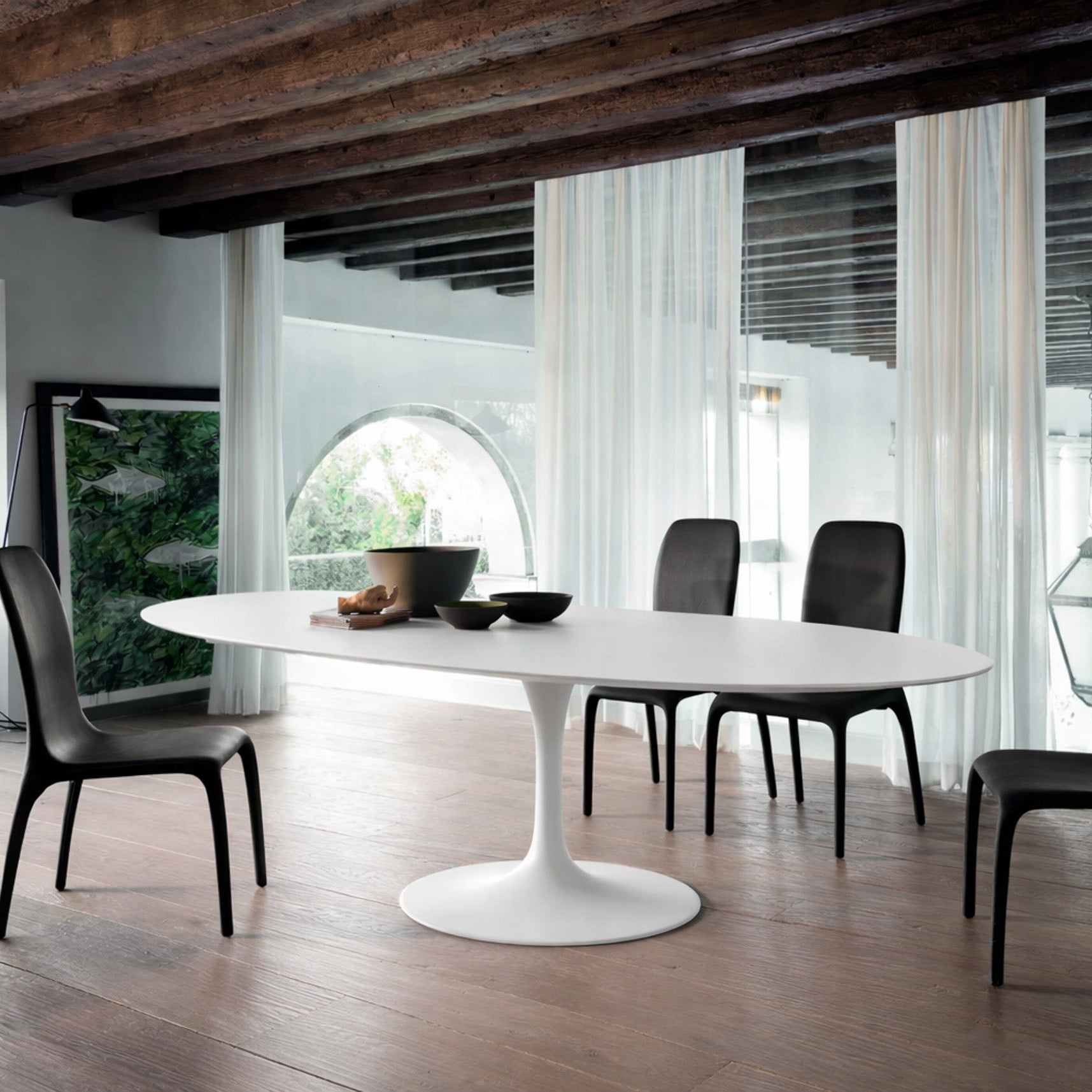 Oval white marble dining table Saarinen style with white pedestal base in a modern architectural interior with exposed wooden beams and arched window, styled with black chairs and sheer curtains