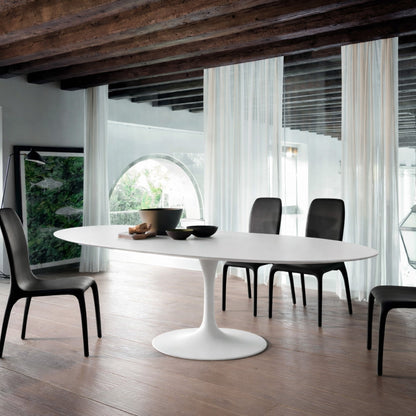 Oval white marble dining table Saarinen style with white pedestal base in a modern architectural interior with exposed wooden beams and arched window, styled with black chairs and sheer curtains
