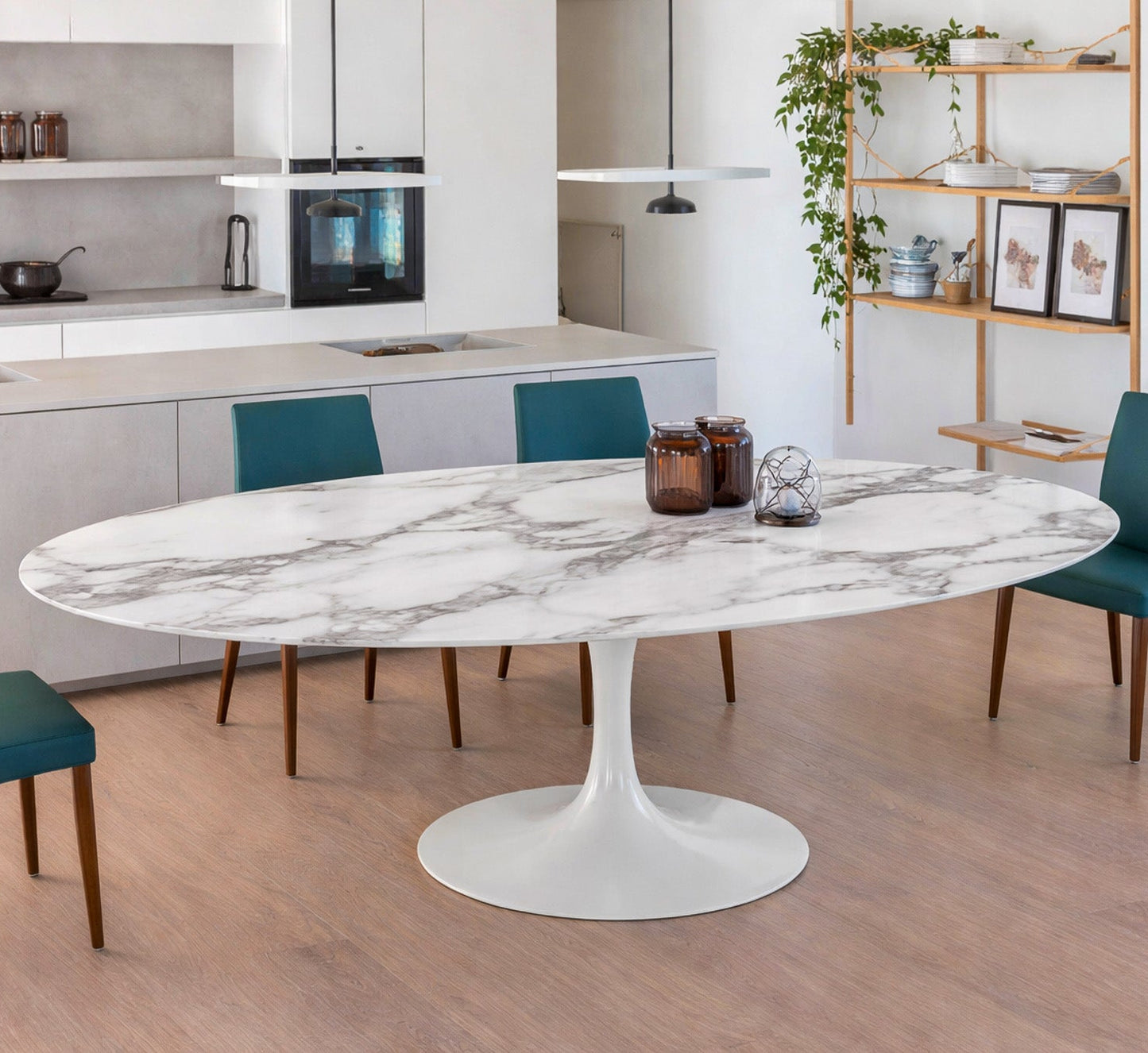 Oval white marble dining table with white pedestal base in a modern kitchen interior, styled with teal upholstered chairs, open shelving and pendant lighting