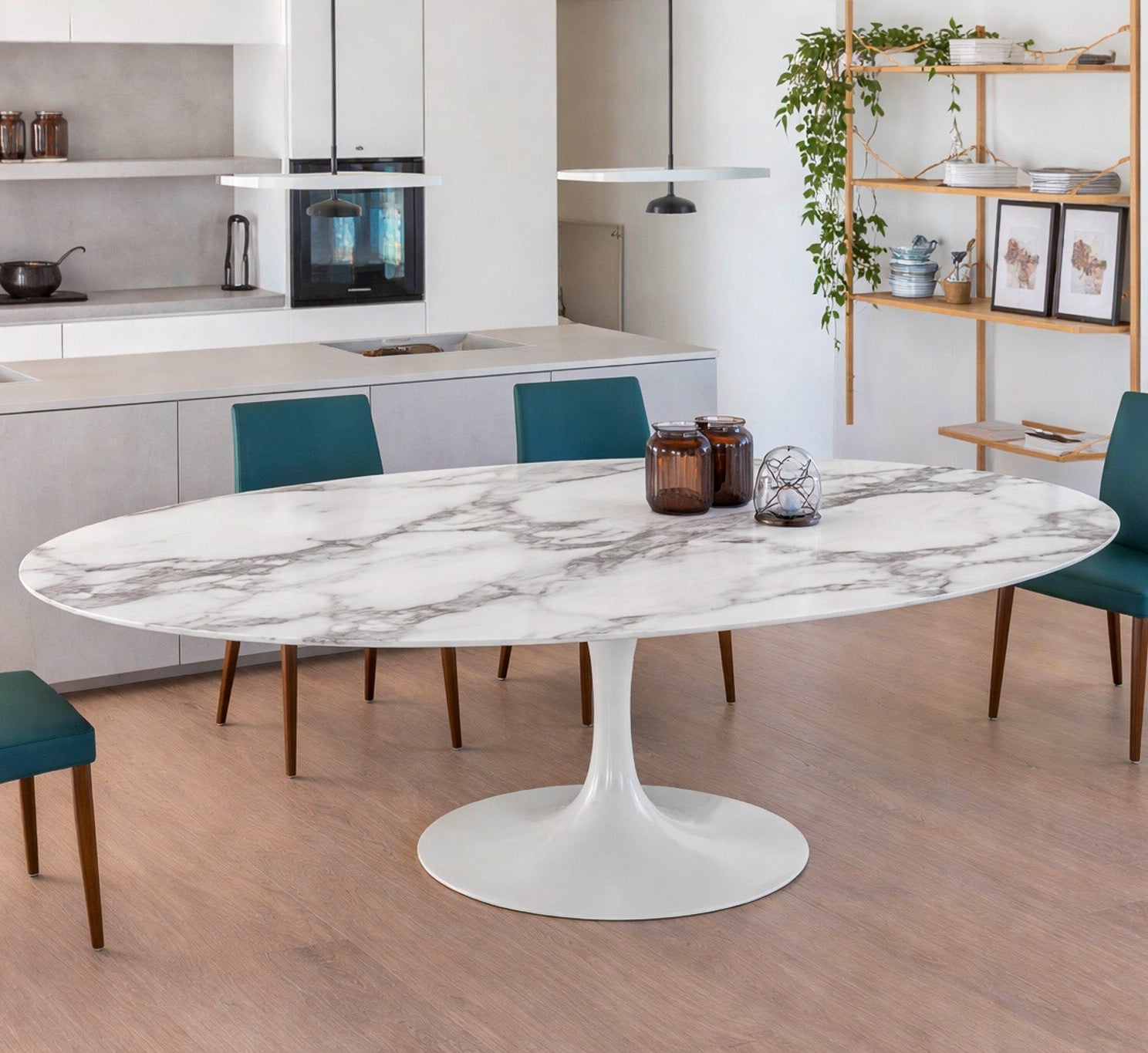 Oval white marble dining table with white pedestal base in a modern kitchen interior, styled with teal upholstered chairs, open shelving and pendant lighting