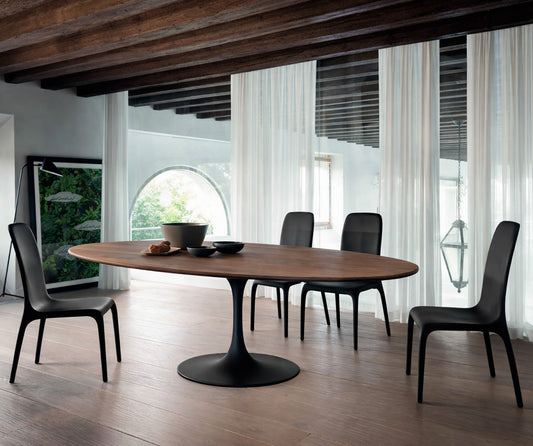 Oval wooden dining table with black pedestal base in a modern architectural interior with exposed wooden beams, sheer curtains and arched window, styled with black chairs