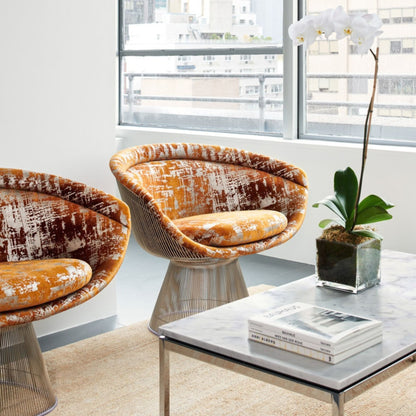 Pair of Platner wire lounge chairs upholstered in orange patterned fabric beside a marble coffee table in a modern living room.