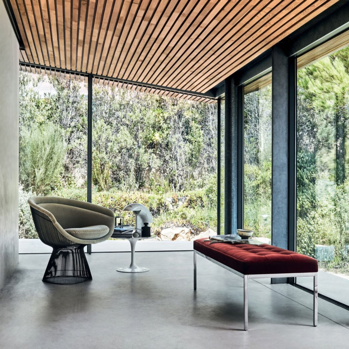 Platner wire lounge chair with light grey upholstery paired with a red upholstered bench in a contemporary glass-walled living room.