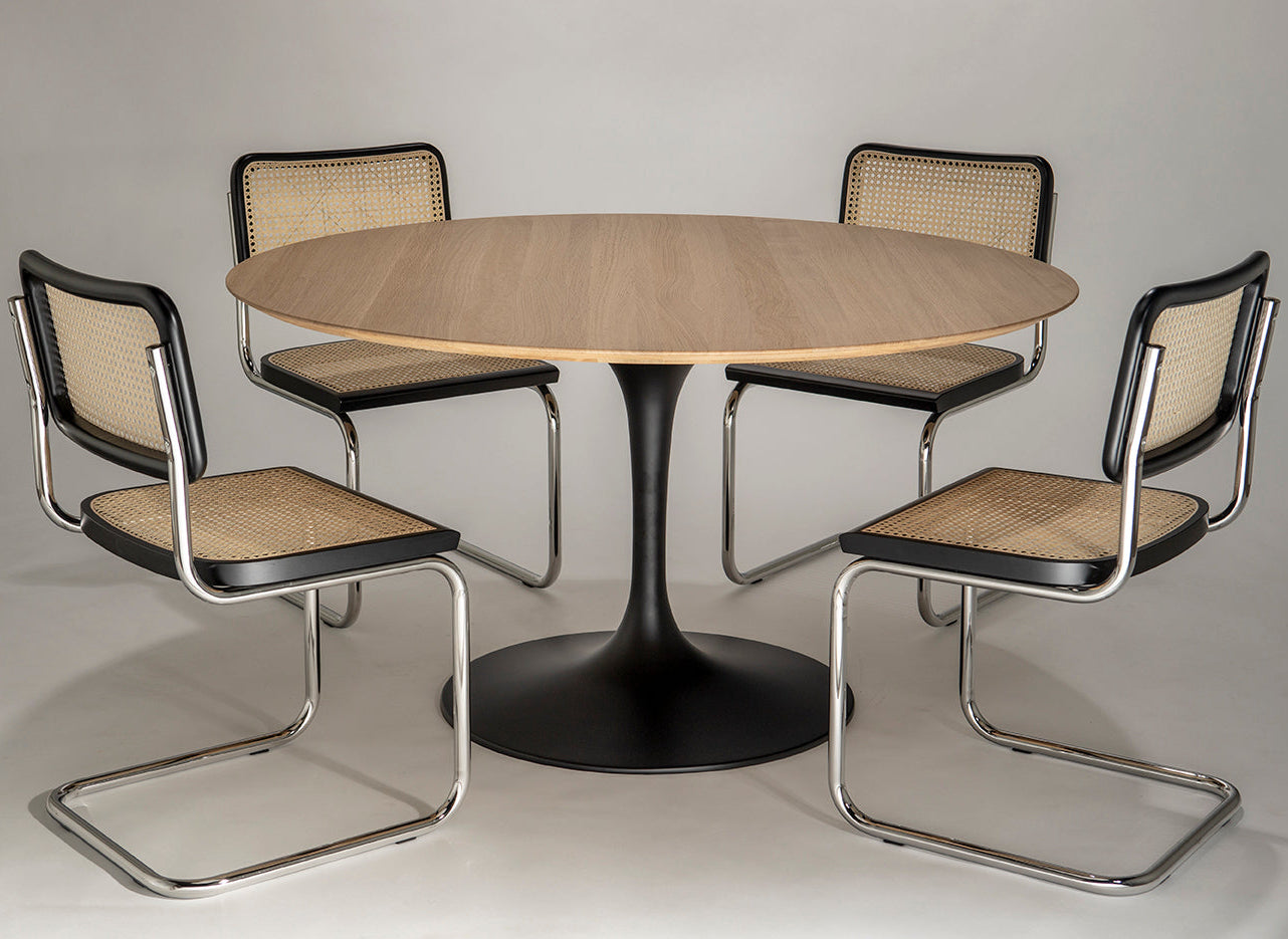 Round oak tulip dining table with black pedestal base paired with four cantilever chairs featuring cane seats and chrome frames on a neutral background
