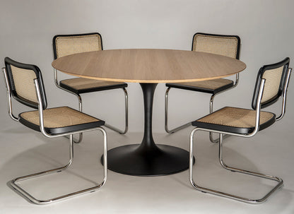 Round oak tulip dining table with black pedestal base paired with four cantilever chairs featuring cane seats and chrome frames on a neutral background
