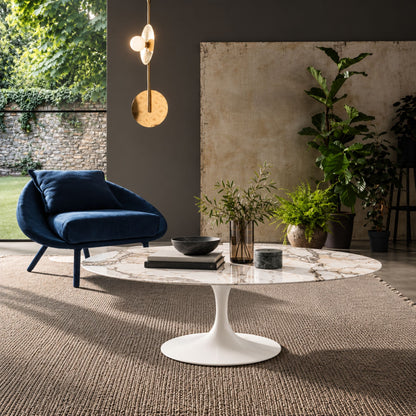 Round tulip coffee table with a white marble-effect top and white pedestal base in a contemporary living room with modern furniture and indoor plants.