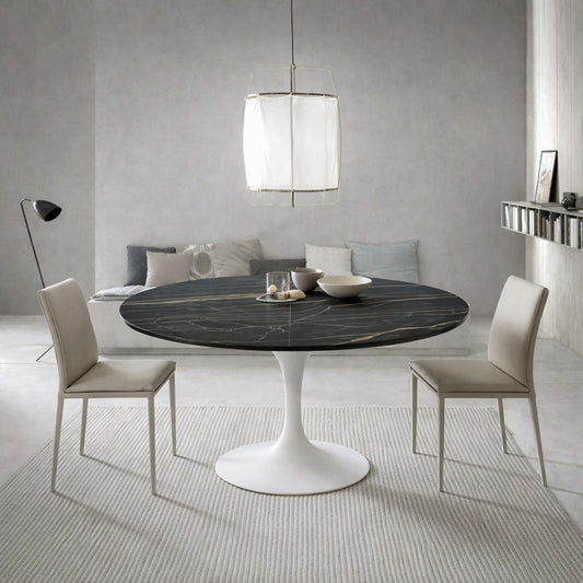 Round tulip dining table with black marble top and white pedestal base paired with beige upholstered chairs in a modern minimalist dining room with neutral tones and soft lighting.