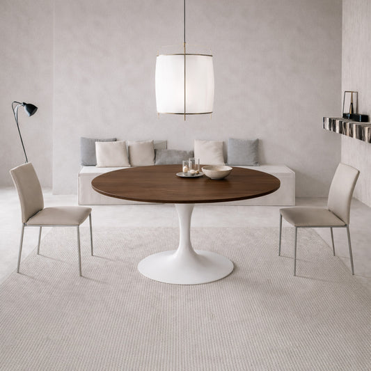 Round walnut tulip dining table with white pedestal base paired with beige upholstered chairs in a modern minimalist dining room with neutral tones and soft lighting.