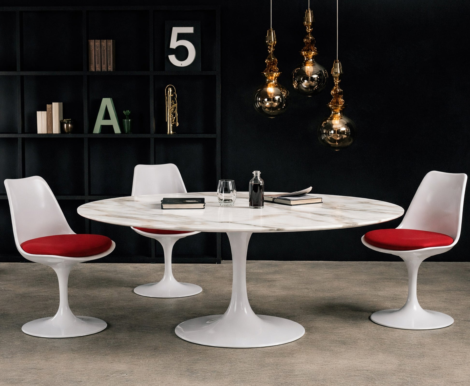 Round white marble dining table with white pedestal base in a dark modern interior, styled with white chairs with red seat cushions, black shelving and glass pendant lighting