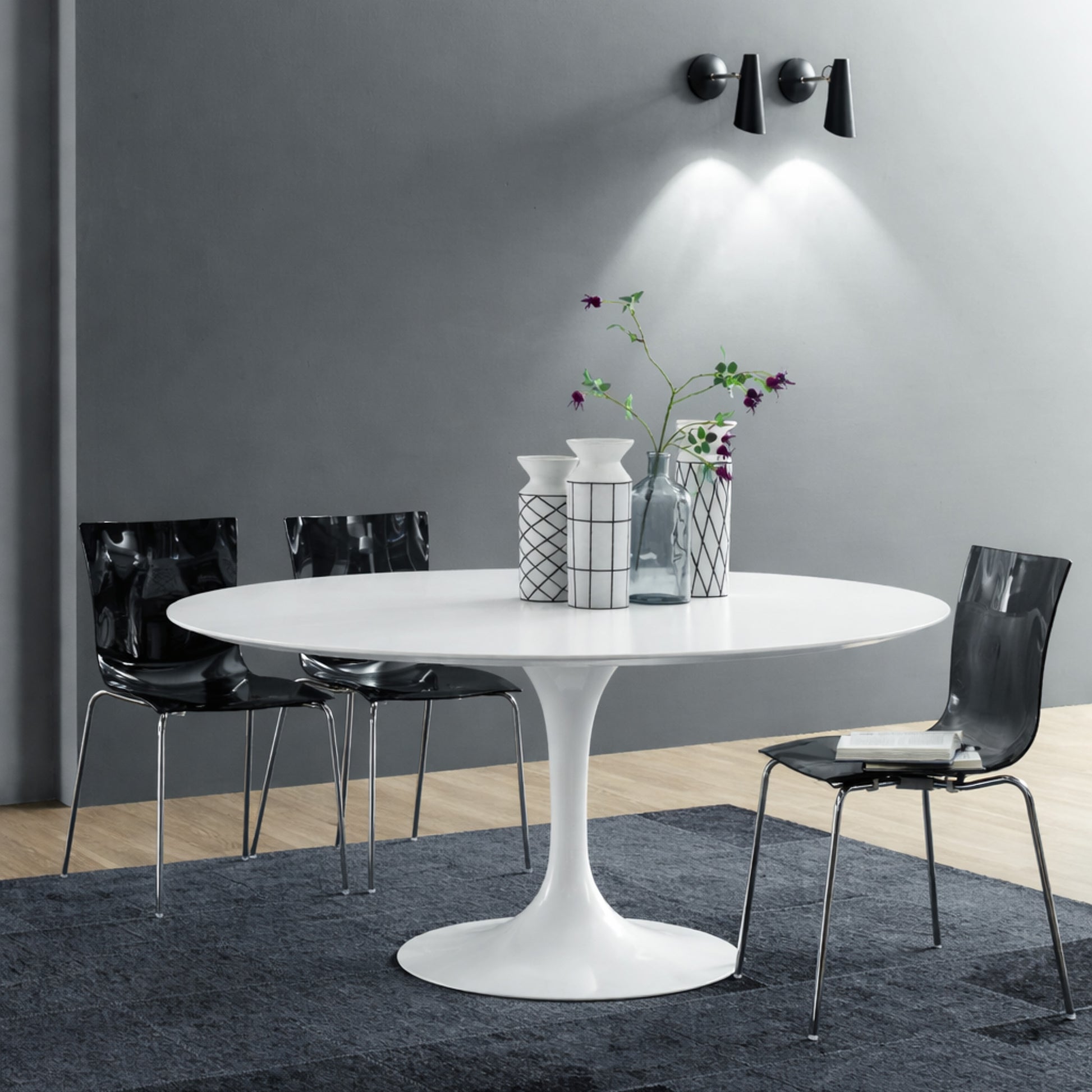 Round white marble dining table with white pedestal base in a modern dark interior, styled with black chairs, grey wall and wall-mounted lighting