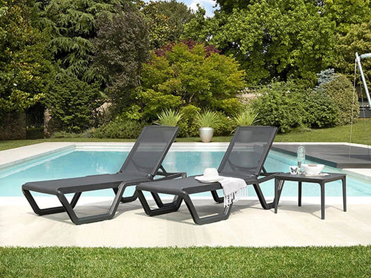 Two modern outdoor sun loungers beside a swimming pool with matching side tables in a landscaped garden.