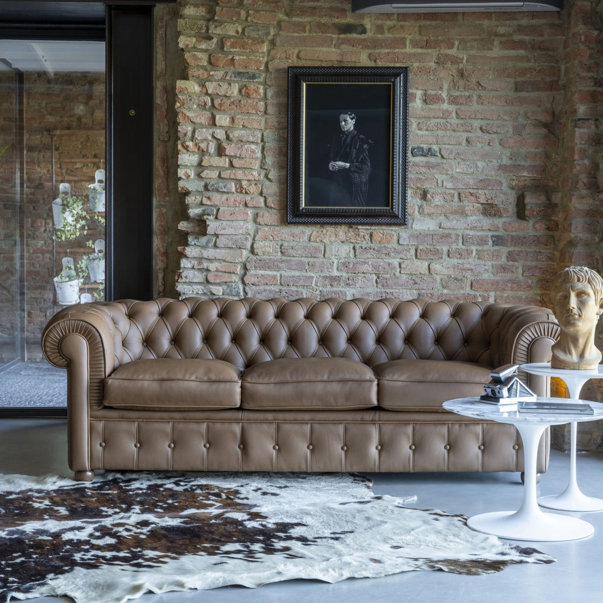 Taupe Chesterfield leather three-seater sofa with deep button tufting, rolled arms, and a timeless British-inspired design, styled in a contemporary loft living room with exposed brick walls