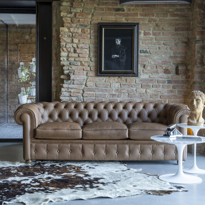 Taupe Chesterfield leather three-seater sofa with deep button tufting, rolled arms, and a timeless British-inspired design, styled in a contemporary loft living room with exposed brick walls